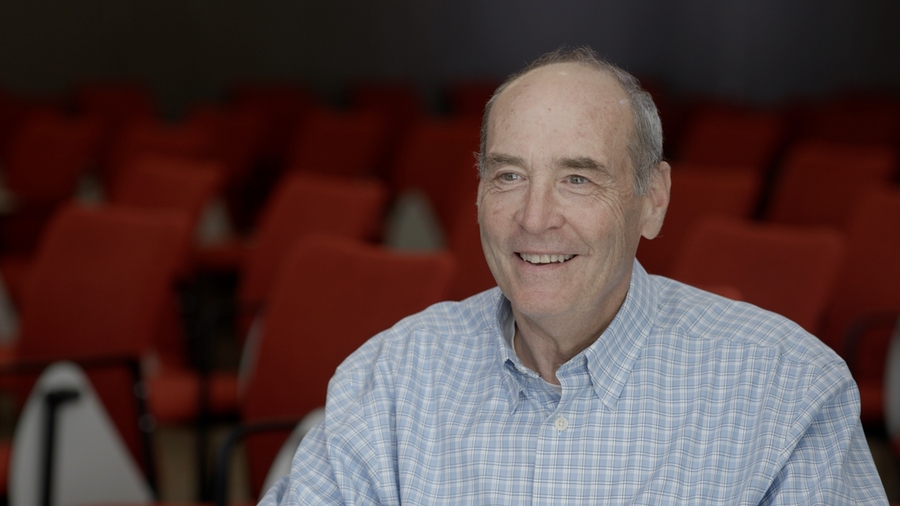 Mark Bear smiles as he is a seated in an auditorium. Rows of unoccupied red chairs are in the background.