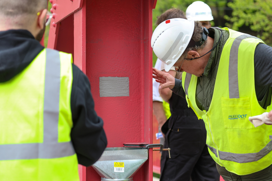 A man wearing a MassDOT safety vest and hardhat examines a gray patch on an orange I-beam