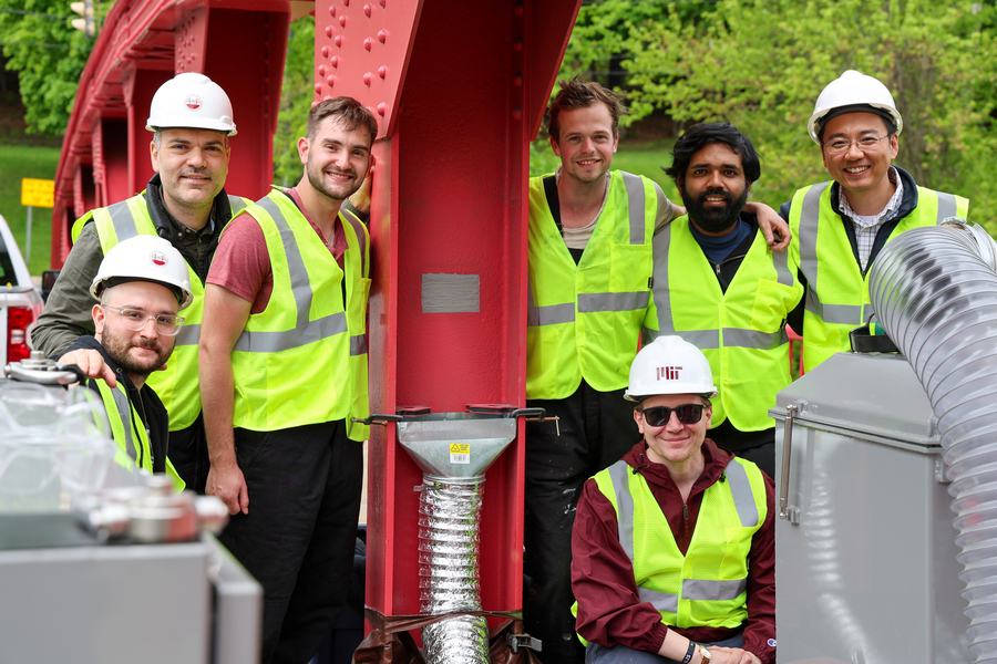 Seven men wearing safety yellow work vests and hardhats pose by a metal I-beam that has a small gray patch in its center.