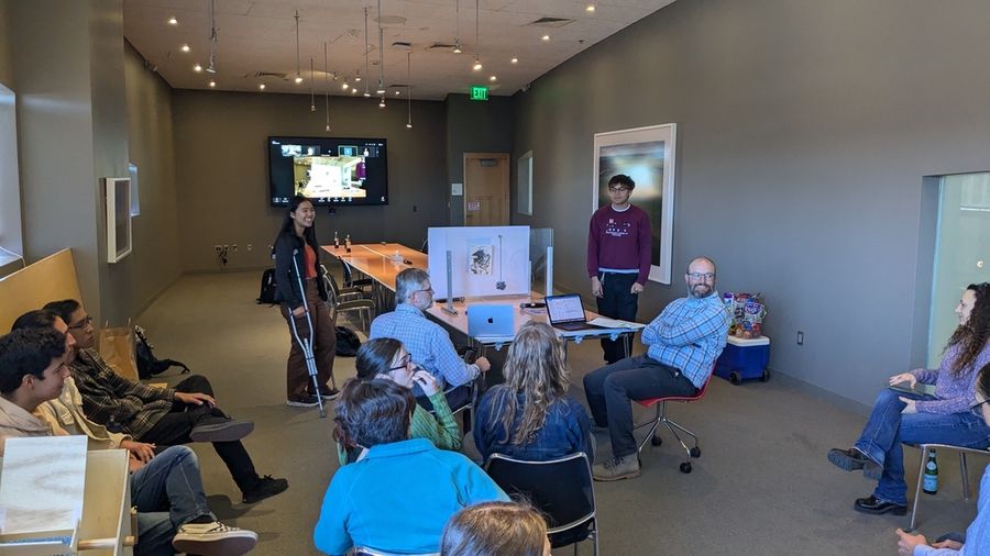 About a dozen students gather in a conference room, presenting their prototypes on a long rectangular table