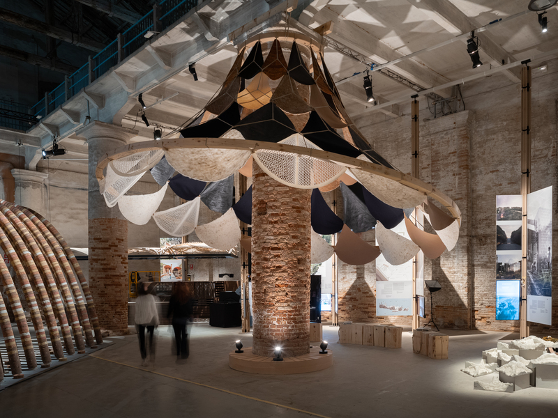 Architectural installation with layered panels of fabric, wood, and bio-based materials suspended from a brick column resembles a giant lampshade.