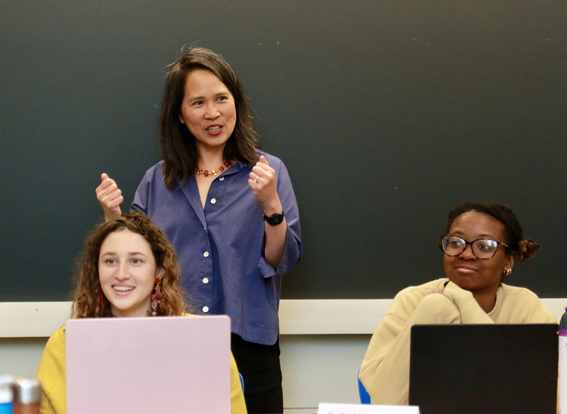 Lily Tsai stands behind Leela Fredlund and Karenna Caton, who are seated at a table with open laptops.