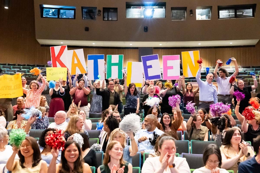 Smiling people stand up from bleacher seats and hold colorful signs that spell “Kathleen.” 