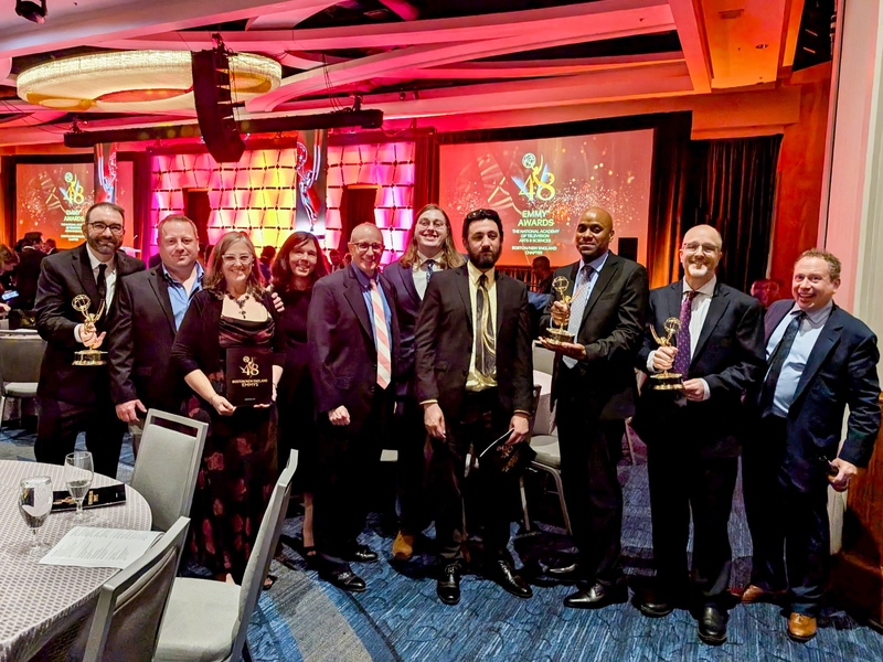 Group photo of 10 adults smiling in formal wear at an awards ceremony. Three are holding Emmy trophies.