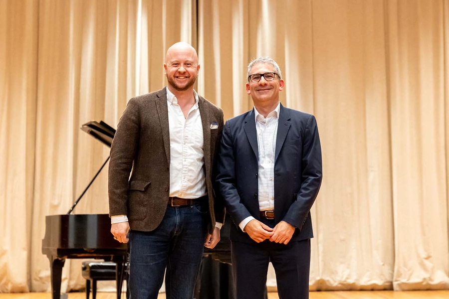 Two men in suits stand next to each other, smiling, on the auditorium stage. 