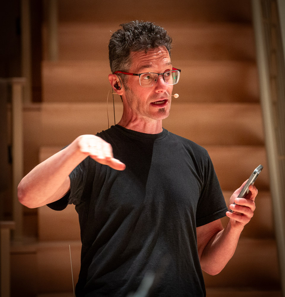 Eran Egozy, wearing head-mounted microphone and holding a cellphone, addresses an unseen audience
