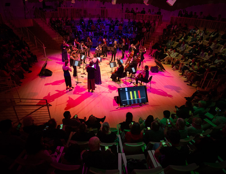 Top-town view of 18 musicians playing on a circular stage under red light, surrounded by an audience