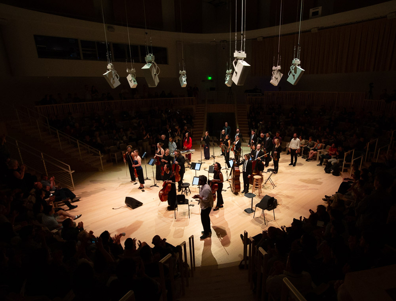 18 musicians stand on a circular stage under white lighting