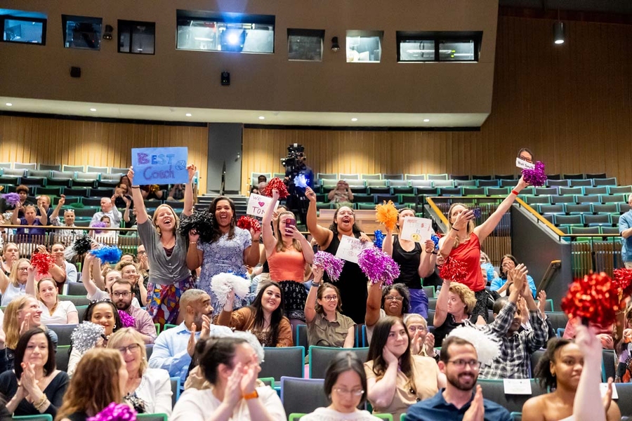 Audience members standing in bleachers wave pompoms, clap, cheer, and hold signs of encouragement.
