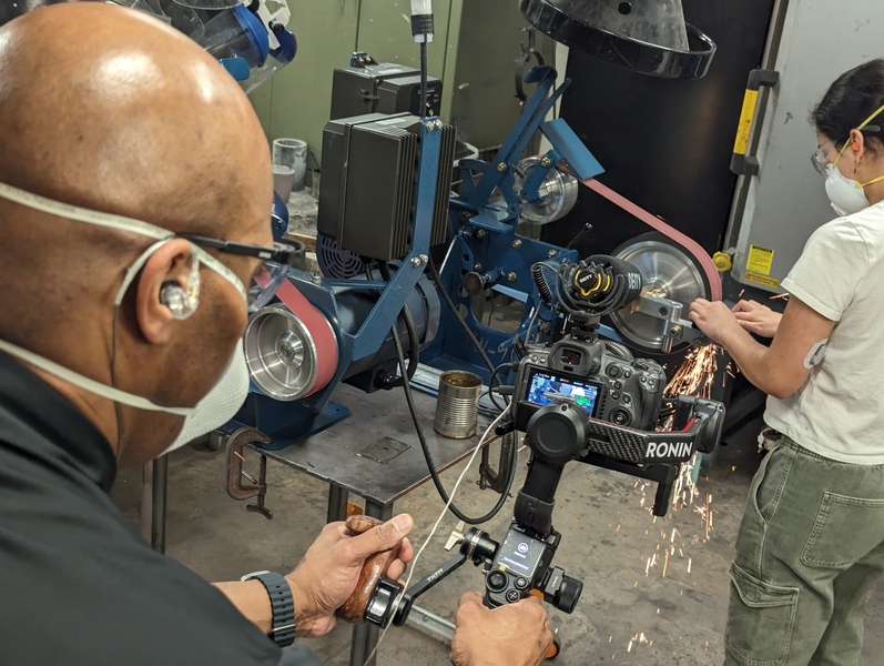 Candid photo of a cameraman filming a college student wearing protective gear who is sharpening a knife on a grindstone.