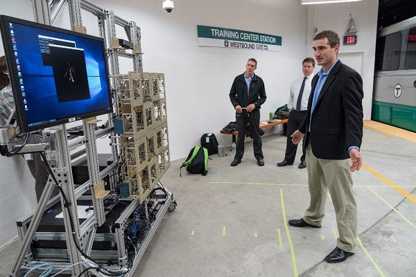 An individual stands in front of an imager as two individuals look on at a subway station platform.