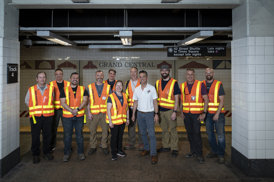 11 people, all but one wearing orange safety vests, pose on a Grand Central subway platform.
