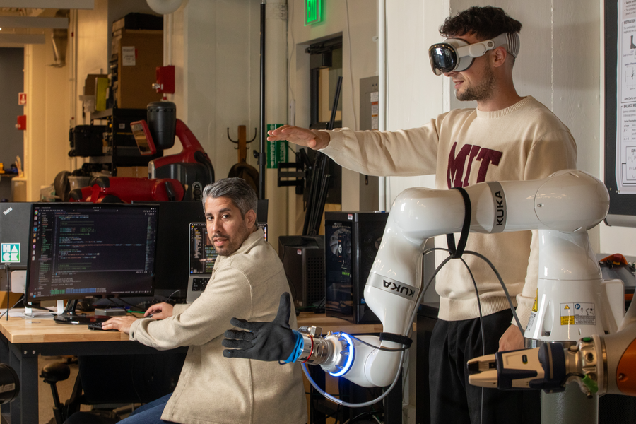 Two researchers, one seated at a keyboard, one standing and wearing VR goggles to interact with a robotic arm
