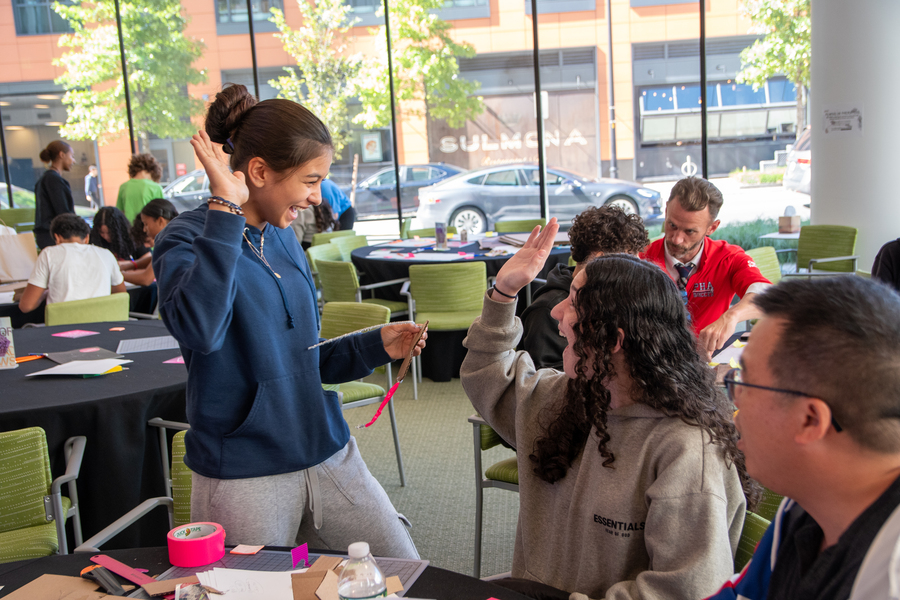 A standing student high-fives a sitting student during a hands-on design activity at MIT, surrounded by tables with craft materials and other participants working in groups.