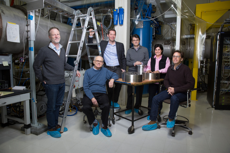 Rai Weiss and five colleagues sit around a table with instrumentation on top, in a lab. They all wear blue slippers over their shoes.