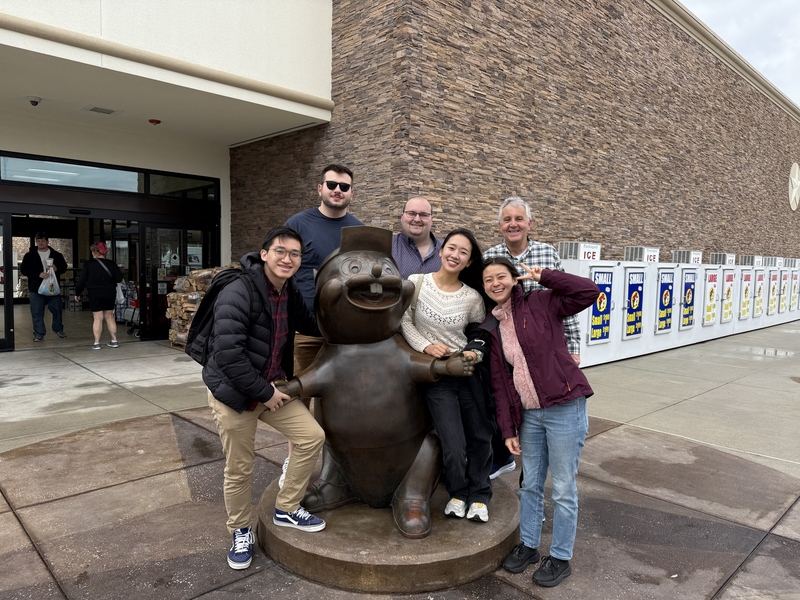 6 people pose with a large bronze statue of a cartoon beaver beside a brick building