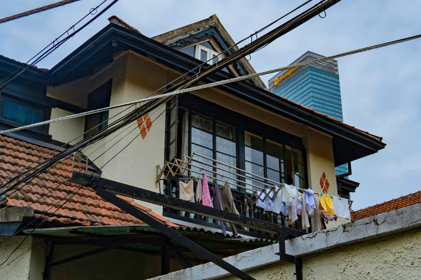 The upper story of a house, with laundry hanging from a balcony. A glass high-rise is visible in the background.