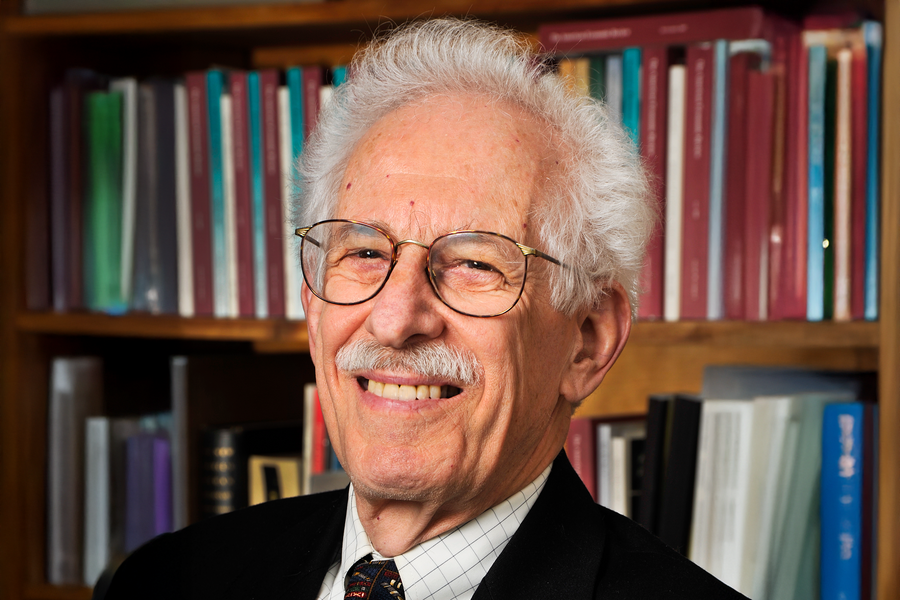 headshot of smiling white-haired man wearing glasses, with bookshelves in the background