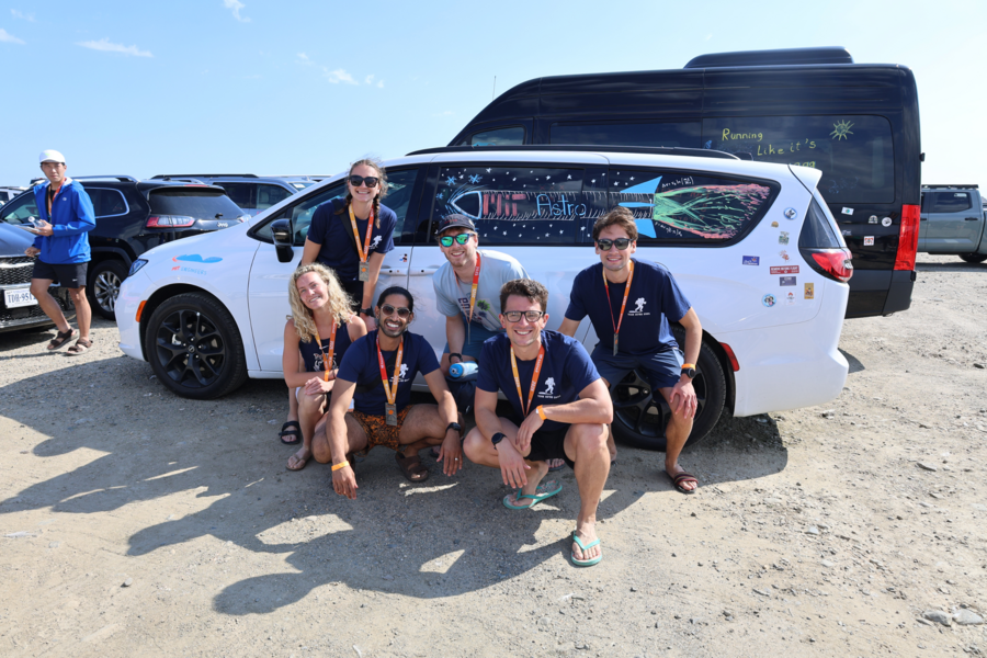 Seven smiling individuals in matching blue T-shirts kneel and stand beside a decorated white van in a sunny parking lot.