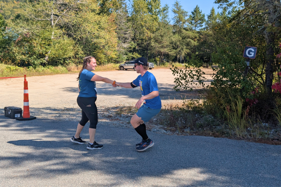 A man and woman exchange a baton during an outdoor relay race on a sunny day. They're in a parking lot surrounded by trees, with a traffic cone nearby.