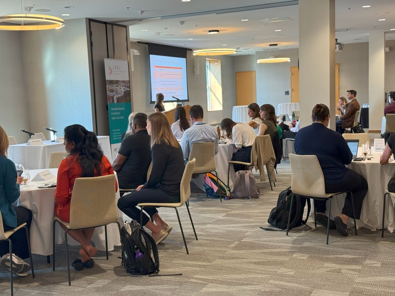 More than 20 people sit at round tables in a conference room watching a presenter speak to slides projected on a screen.