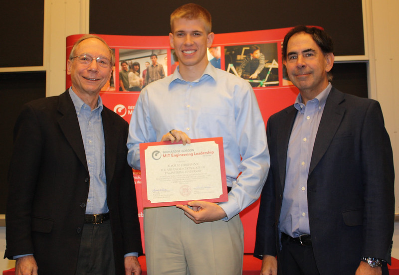 Adam Fuhrmann stands holding a certificate. He's flanked by Joel Schindall and Ed Crawley.