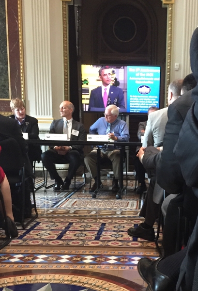 Gregory Olson and other panelists sit at table under a monitor featuring Barack Obama at the White House.
