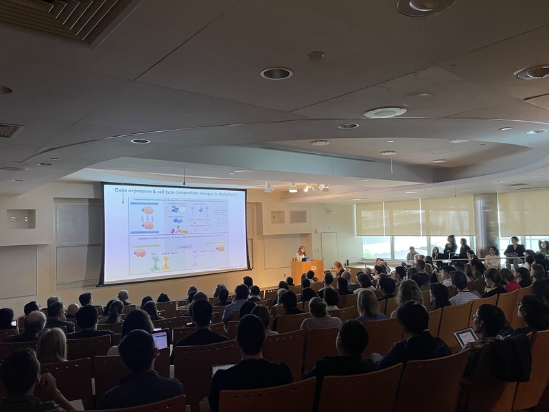 A wide shot of Singleton Auditorium in MIT's Building 46 shows an audience that fills the seats while a speaker at the front of the room presents an Alzheimer's research slide on a large screen