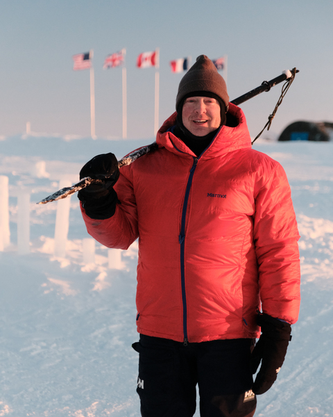 David Whelihan, wearing a red parka, stands on ice in the Arctic with a walking stick over his shoulder; flags representing different nations are pictured in the background.