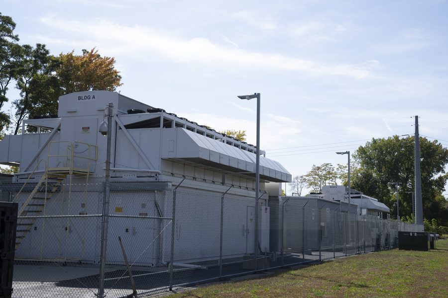 An exterior shot of a data center pod: two blocky, utilitarian gray buildings are visible inside a chain link fence.
