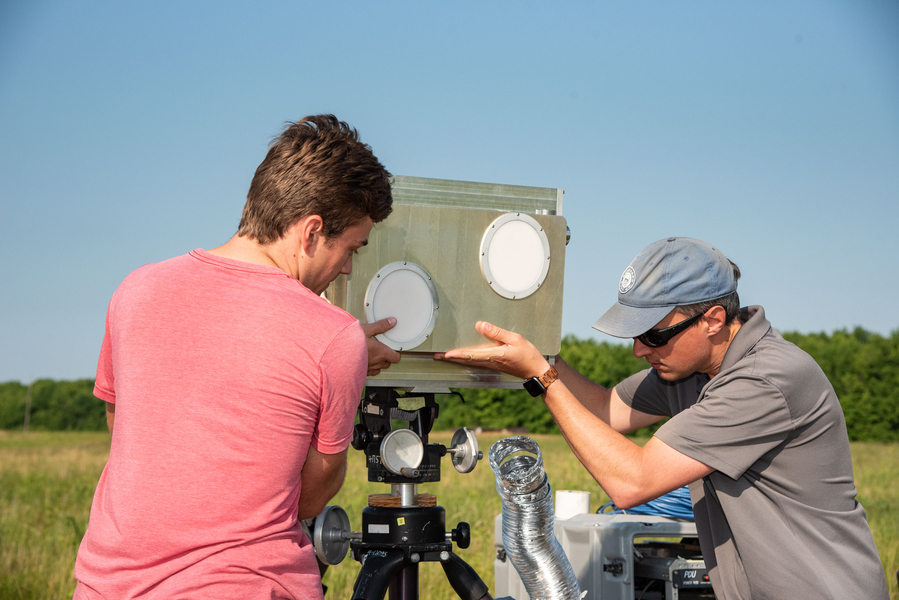 Two men adjust a metal antenna unit, approximately 12 in by 18 in, on a tripod in a field.