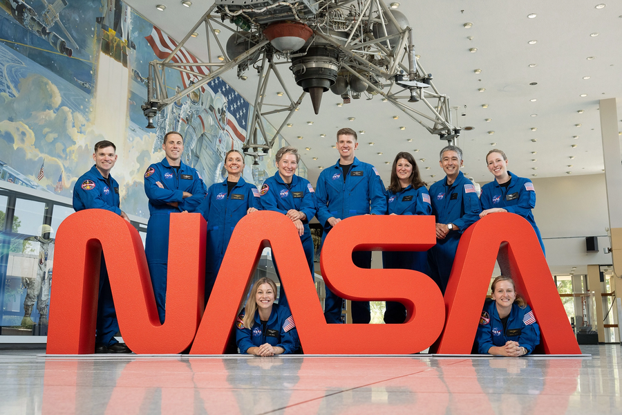 10 astronaut candidates in blue pilot suits flank a large red NASA statue inside a vestibule with an old lunar module hanging from the ceiling