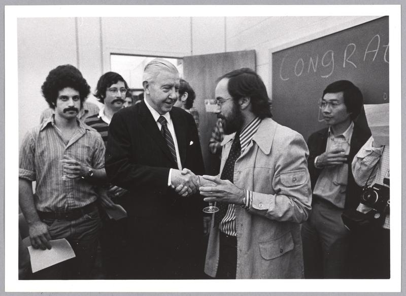 Greyscale photo of some 10 people in a small room, holding glasses, as James Killian shakes David Baltimore's hand. A chalkboard on the right has "Congratulations" on it.