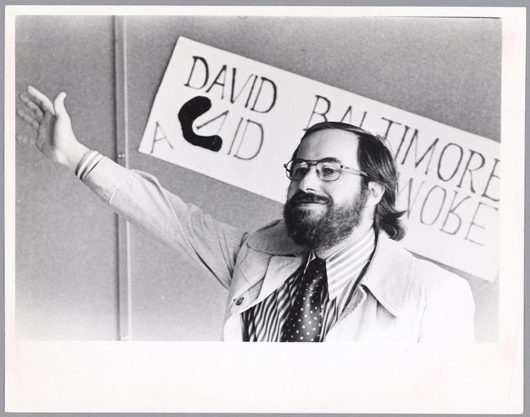Greyscale photo of David Baltimore raising one arm while standing before a sign that says "David Baltimore" twice, once right-side-up, and once upside-down like a mirror image. On the upside-down name is a dark blob that surrounds the "V" and appears to have displaced the "D" and "A."