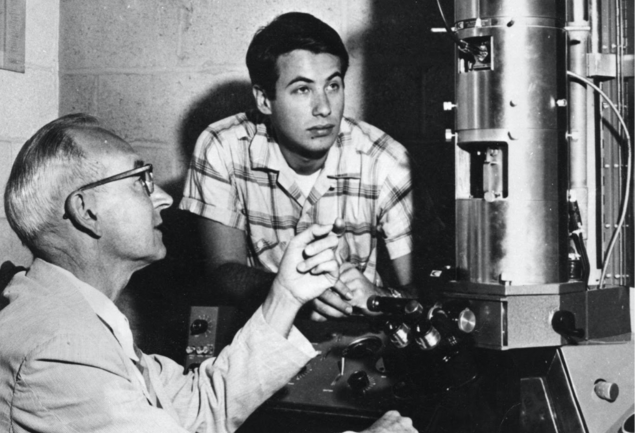 Greyscale photo of an older man operating large scientific equipment while a young David Baltimore watches intently.