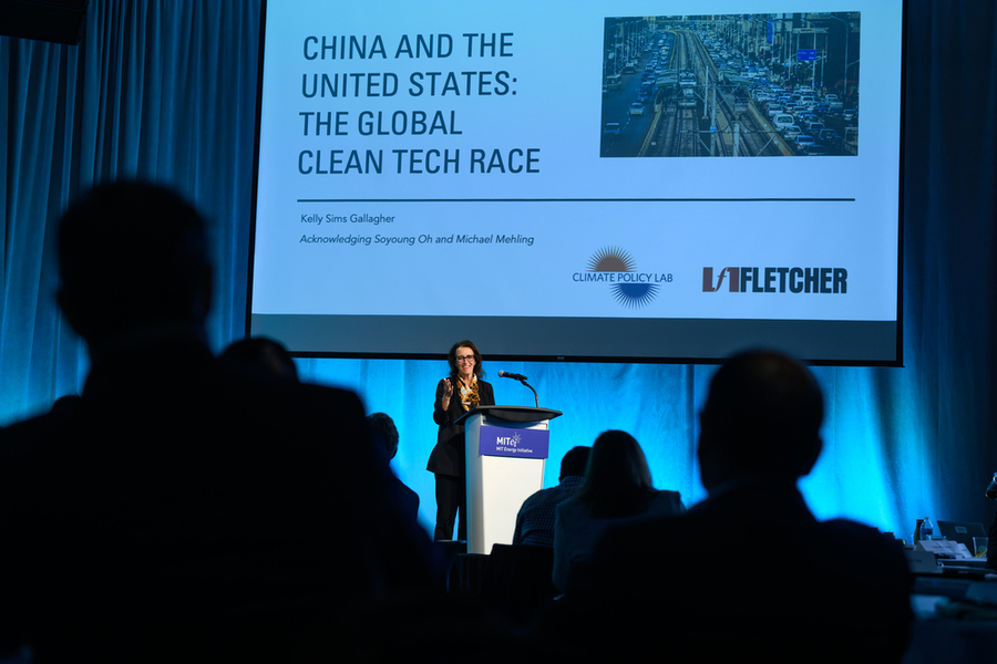 Speaker stands at a lectern under a large projected slide with an audience in the foreground.
