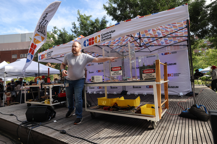 Joshua Wolfe stands on a small outdoor stage, pointing at some equipment labeled "danger high voltage"