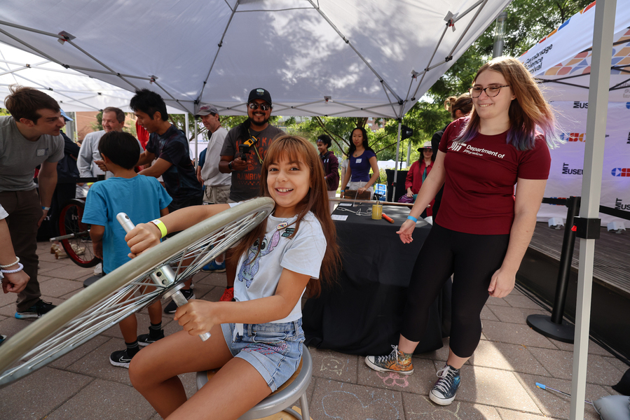 Girl holds onto a spinning bike wheel under a small tent with others looking on