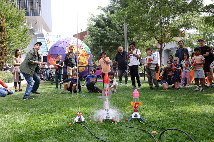 3 small water rockets at various stages of liftoff on a grassy area as about 20 kids and adults look on