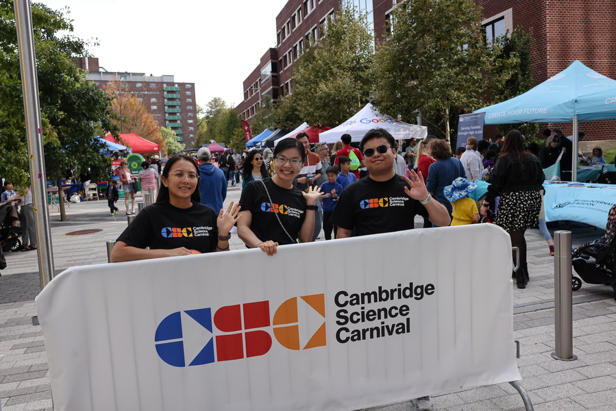 Three young adults wave to the camera in front of a Cambridge Science Carnival banner. Various tents are behind them with people milling about.