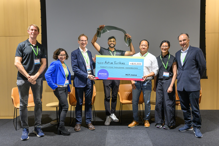 A man poses behind a giant check with his invention held overhead. Flanking him are two people holding the check, and four other people flanking them.
