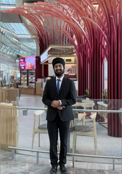 Amitoj Singh in a black suit and turban and a blue tie stands in front of a cafe with red decorative structures and a glass ceiling.