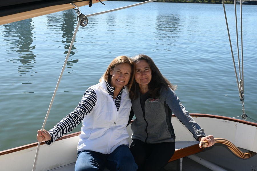 Carrie Moore and Jennifer Light sitting with an arm around one another in a sailboat on the Charles River.