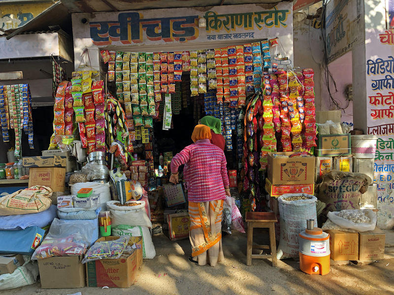 Micro-grocery store in India with items for sale in boxes and hanging out front and two people with their backs to the camera