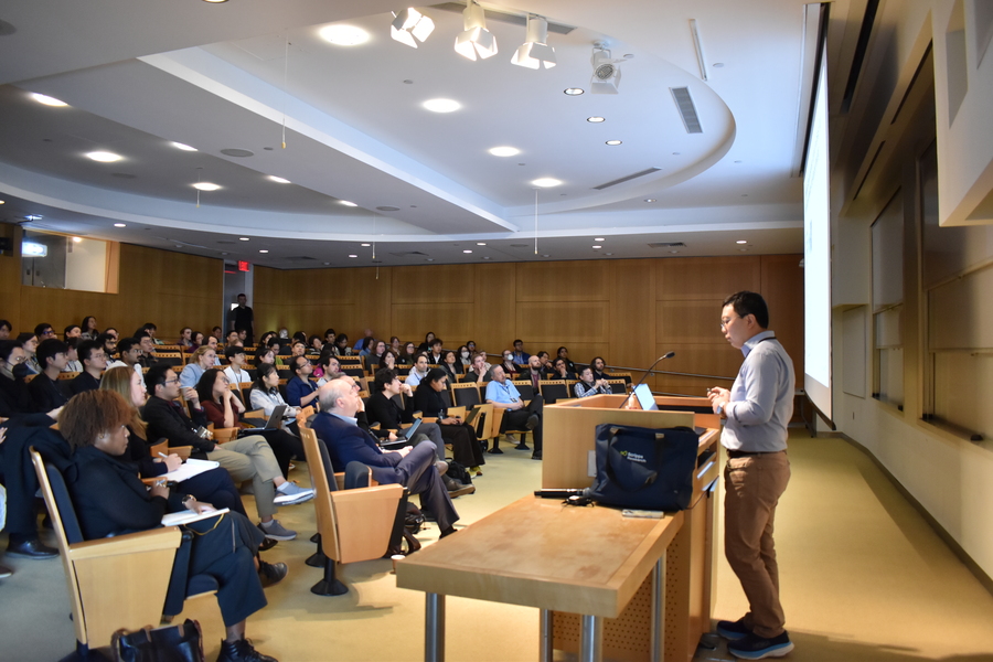 Side view of Li Ye, standing behind a lectern, speaking to a near-full audience at MIT's Singleton Auditorium