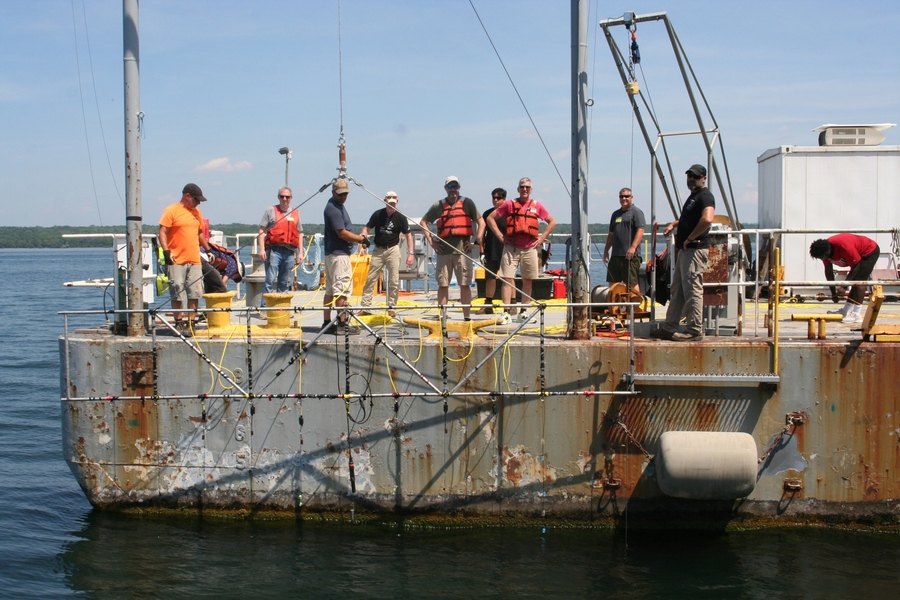 10 people stand on a rusty barge in Seneca Lake, NY, lowering a metal frame into the water.