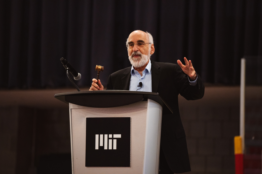 John Sterman speaking at a lectern, gesturing with one hand and holding a small gavel in the other