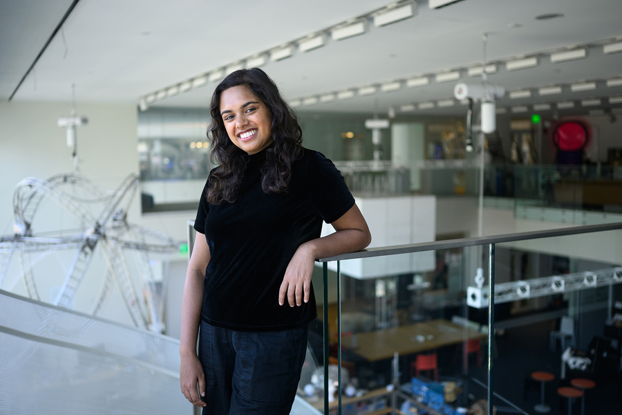 Kimaya Lecamwasam poses on an atrium balcony, with lab spaces behind her and a sculpture resembling a circular roller coaster hanging from the ceiling