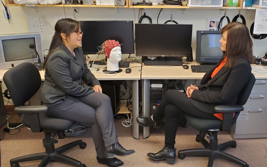 Diane Chan and Li-Huei Tsai sit, talking, at a table covered in research equipment. In front of powered-off monitors sits a polystyrene head with a red cap of electrodes