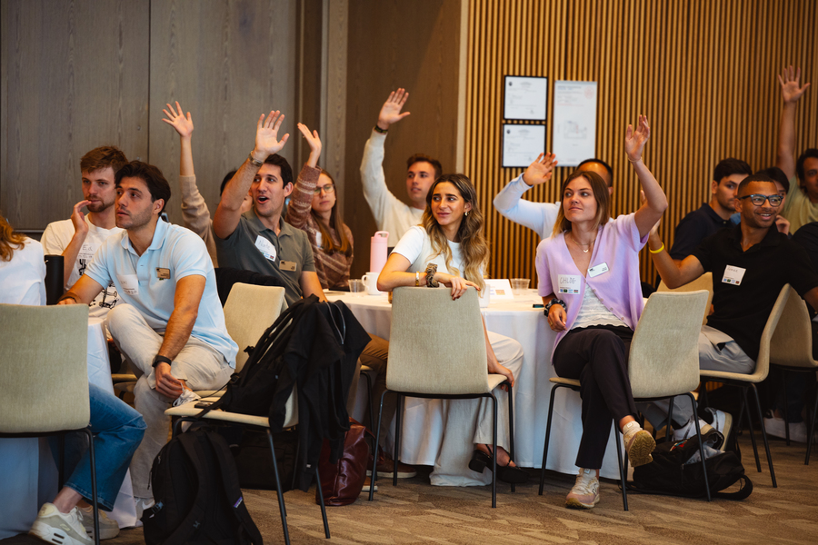 About a dozen students are pictured sitting at round tables, half of them raising their hands